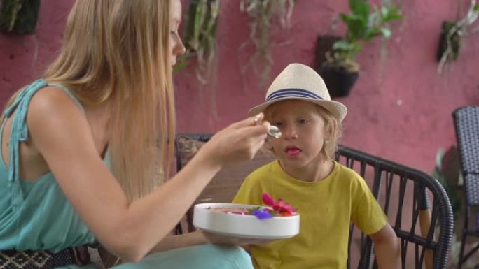 年轻女子和她的小儿子坐在一个美丽的咖啡馆里，在碗里享受五颜六色的格兰诺拉麦片冰沙。健康饮食理念。慢动