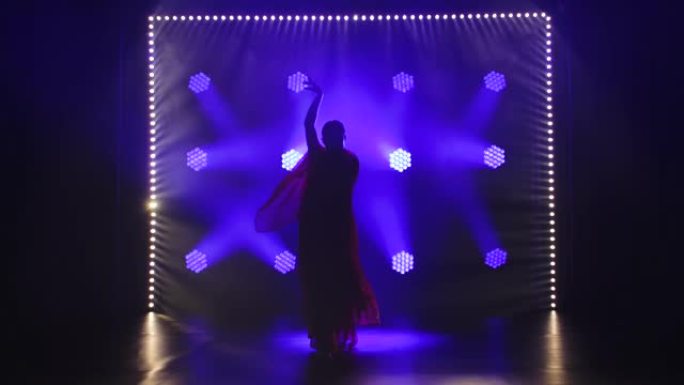 剪影一个穿着红色纱丽的年轻女孩舞者。印度民间舞蹈。在带有烟雾和蓝色霓虹灯的黑暗工作室中拍摄。慢动作