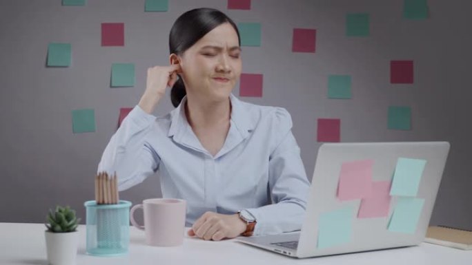 亚洲妇女在笔记本电脑上工作，发痒，并将手指插入她的耳朵坐在家庭办公室。