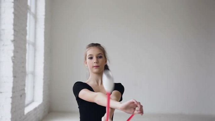 艺术优美的体操运动员手持马鞭子，在体操大厅的白色背景上进行杂技动作。女孩穿着黑色的身体和粉红色的裙子