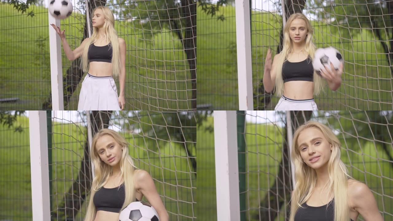 自信美女在足球网与球合影的肖像。摄像机接近站在户外操场上微笑的苗条白人女运动员。女子足球运动员在户外