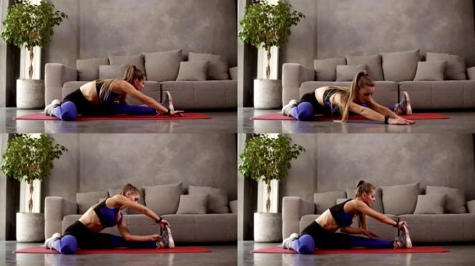 穿着运动服的长发金发女子在家训练坐在沙发前的垫子上，伸展腿部肌肉，而坐在垫子上的地板上。阁楼内部房间