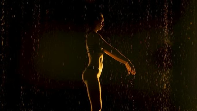 年轻女子在雨中在黑暗的工作室里练习瑜伽体式。黑色背景和黄色霓虹灯。水滴中身体的轮廓。慢动作