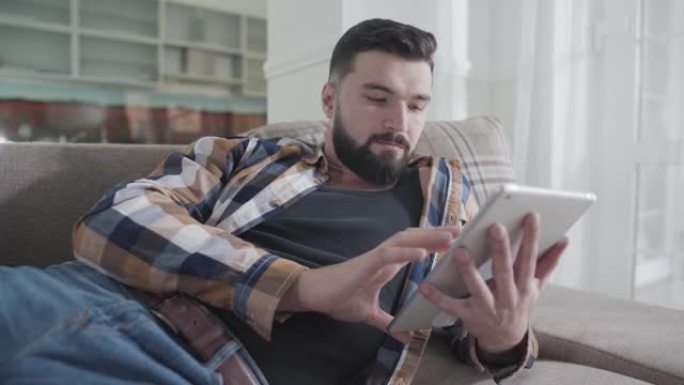 集中的高加索大胡子男子躺在沙发上并使用平板电脑的特写肖像。放松的家伙在家上网。社交媒体成瘾，休闲，生