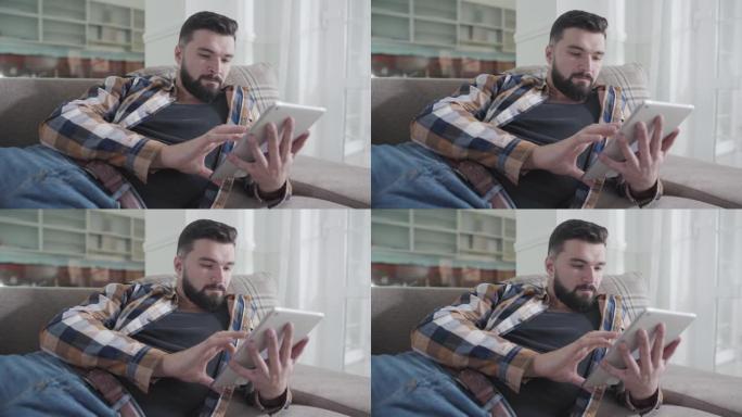 集中的高加索大胡子男子躺在沙发上并使用平板电脑的特写肖像。放松的家伙在家上网。社交媒体成瘾，休闲，生