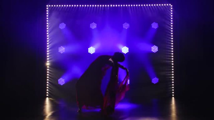 剪影一个穿着红色纱丽的年轻女孩舞者。印度民间舞蹈。在带有烟雾和蓝色霓虹灯的黑暗工作室中拍摄