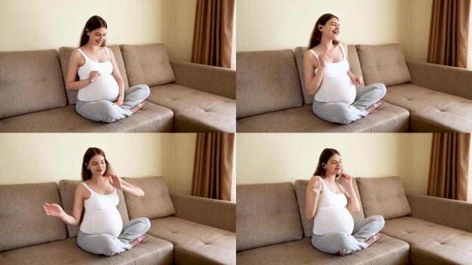 孕妇在沙发上戴耳机。准妈妈在轻便的卧室听音乐跳舞。怀孕的女性在家听音乐