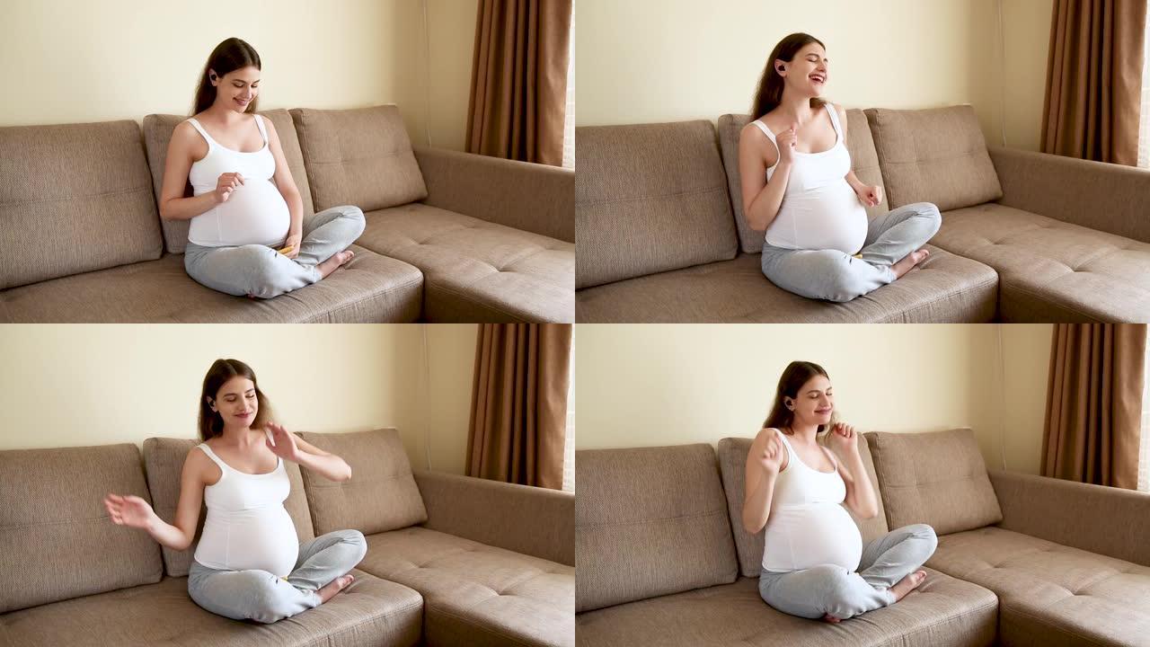 孕妇在沙发上戴耳机。准妈妈在轻便的卧室听音乐跳舞。怀孕的女性在家听音乐