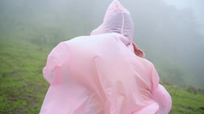 穿着粉色雨衣的年轻亚洲女性徒步旅行者的慢动作后视，在山中刮起大风，在雨中徒步旅行。漂亮女孩在美丽的大