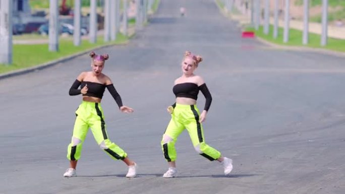 两个穿着运动服的年轻女孩正在跳嘻哈，在宽阔的街道上表演现代青年舞蹈的壮观元素。慢动作