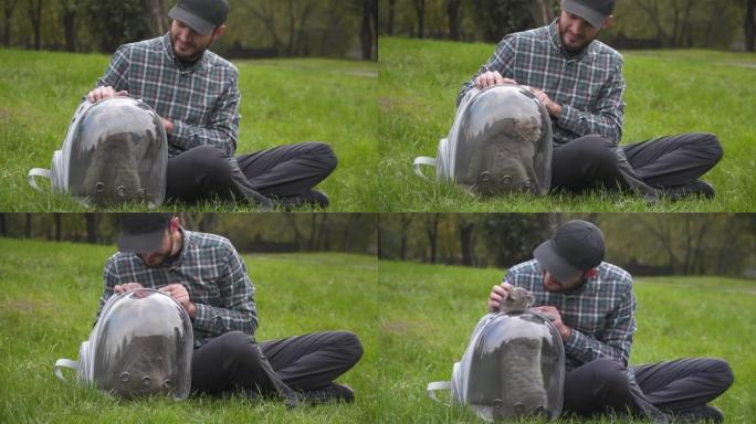 公园里户外有花哨宠物载体的人坐在草坪上，和宠物玩耍。有趣的猫在透明的宠物背包里看起来从背包里出来。带