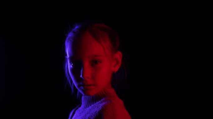 微笑的女孩转过身，在黑暗的工作室里看着相机，红色和蓝色的灯光。美丽的女孩在蓝色和红色背光的深色背景下