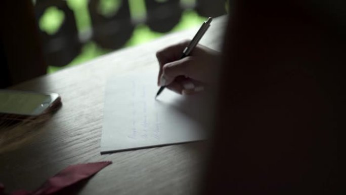 年轻女子用笔在白纸上写给她心爱的新郎的情书，坐在阳台附近房间的桌子上。手机和小麦花束就在附近。婚礼准