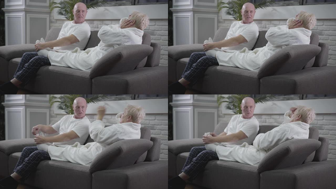 白人老人坐在沙发上按摩妻子的脚的肖像。丈夫照顾他可爱的配偶。快乐的老年夫妇在家休息。永恒的爱，关怀，