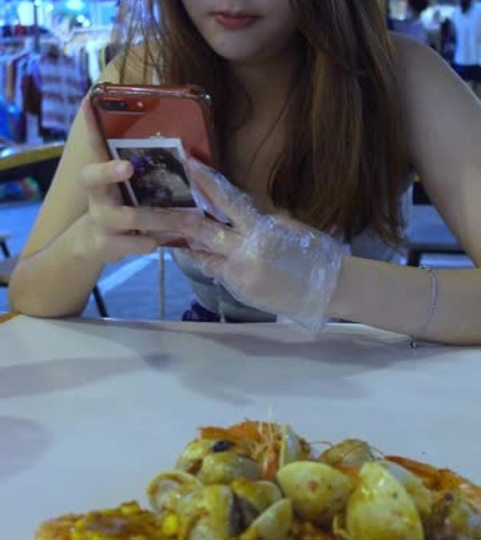 年轻女子在夜市吃街头食品，并使用智能手机拍摄食物照片，垂直格式视频