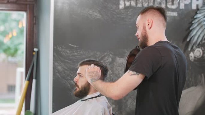 男性理发师手里拿着镜子，留在客户后面，向年轻人展示他的工作成果，他正坐在理发师的椅子上，披着斗篷。窗