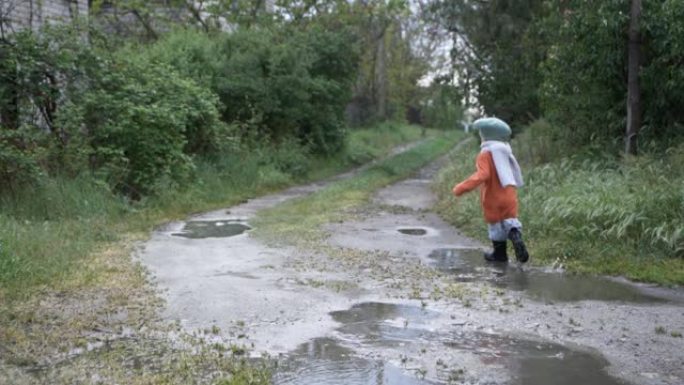 雨后绿树丛中，活跃的快乐男孩戴着帽子和橡胶靴穿过水坑，后视图