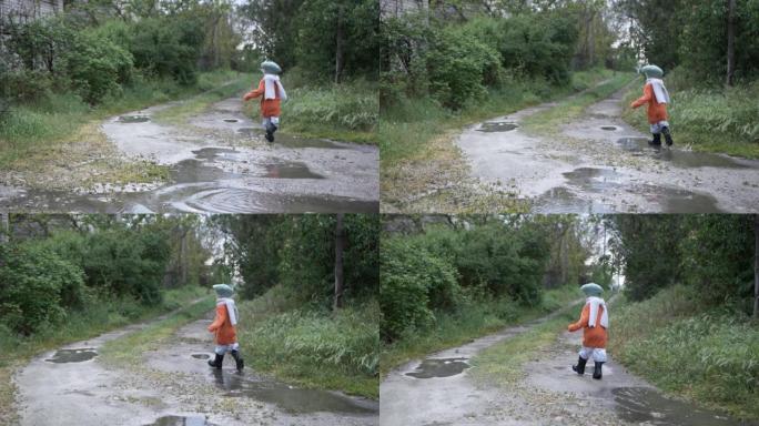 雨后绿树丛中，活跃的快乐男孩戴着帽子和橡胶靴穿过水坑，后视图