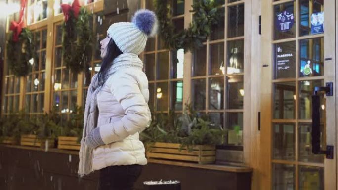 穿着时髦的冬装的积极女孩站在咖啡屋附近，抬头看着雪。女士欣赏圣诞节的节日窗户和街道。