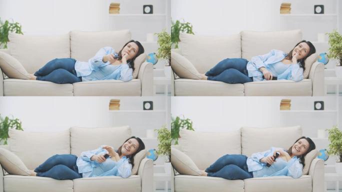 年轻微笑的女人正在用zapper切换频道。她正在看电视，躺在家里的沙发上。复制空间。4K.慢动作。
