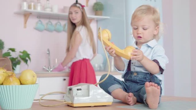迷人的高加索小男孩坐在桌子上玩旋转电话，模糊的年轻女子在后台洗碗。厨房里快乐20世纪60年代高加索家