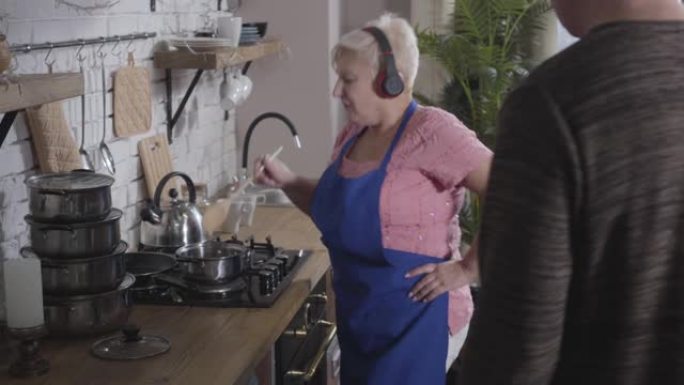 戴着耳机的高加索高级女士在厨房唱歌跳舞。她的丈夫试图和他的妻子说话。快乐的退休人员享受退休后的生活。
