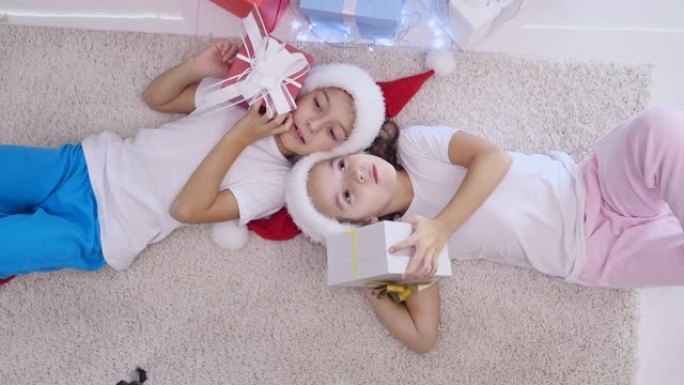 顶视图视频可爱的孩子躺在白色地毯上，摇晃，敲门，听圣诞礼物里面的东西。