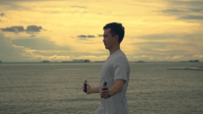 年轻的亚洲运动员穿着运动服跳绳，在夏天美丽的日落时在海滩上锻炼。