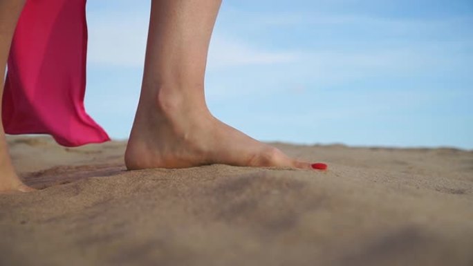 沿着海滩走的苗条女性脚的低侧视。穿着红色连衣裙的面目全非的女人慢慢踩着沙子享受在度假胜地休息。年轻的