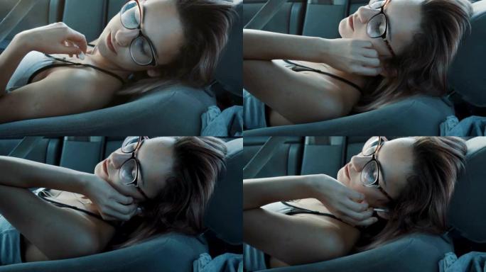一个戴眼镜的女孩坐在前排座位上的汽车里，戴着耳机睡着了。慢动作。