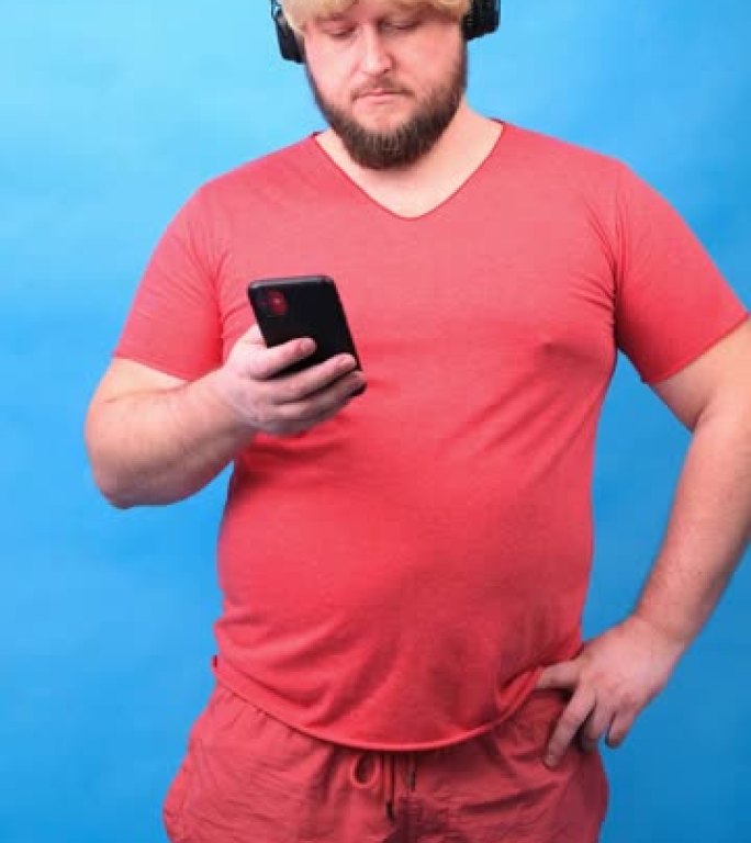 戴着耳机和戴着粉色t恤的假发的滑稽悲伤胖乎乎的怪胎男人看着蓝色背景上的智能手机，垂直