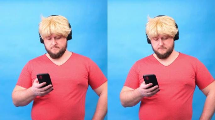 戴着耳机和戴着粉色t恤的假发的滑稽悲伤胖乎乎的怪胎男人看着蓝色背景上的智能手机，垂直