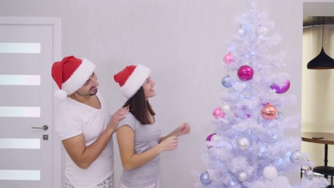 可爱的夫妇看着美丽的圣诞树，女人重新调整粉色玻璃球。