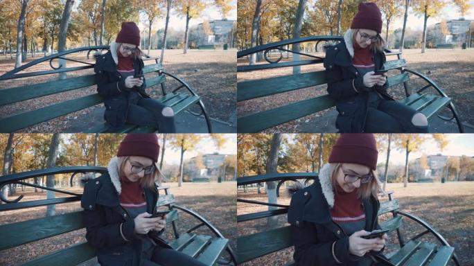 女孩拿着你的手机坐在公园的长椅上。穿着夹克和帽子的女孩