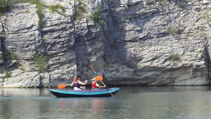 幸福的母子俩在风景如画的岩石和绿色植被的背景下，在一个大山湖水上的充气皮划艇上划船。家庭运动假日山