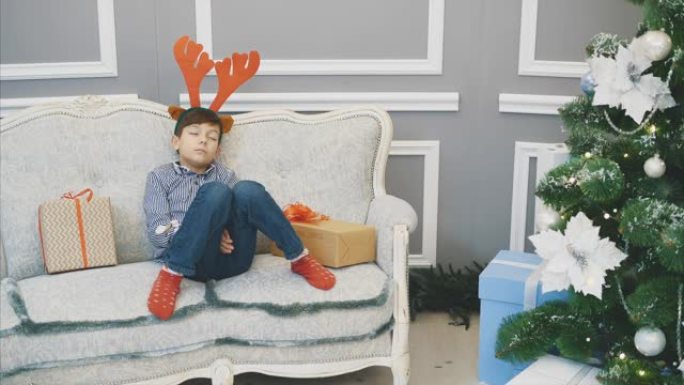 全身可爱的视频，小男孩带着驯鹿头饰，在枞树附近的圣诞节礼物中睡在沙发上。