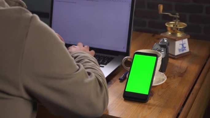 带有绿色屏幕的模型，用于跟踪特写。成年白人男子坐在木桌旁，在笔记本电脑上输入文字。附近有一部带触摸屏