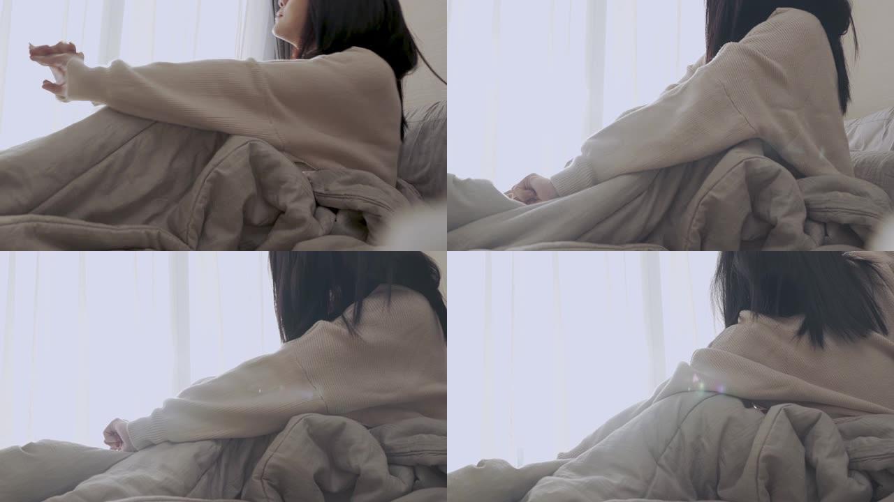 昏昏欲睡的年轻亚洲女性早上刚坐在卧室里的床上，侧视，懒惰的日子凌乱的毯子床白色的窗帘，自然的日光从窗