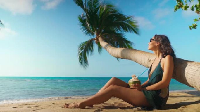 美丽的年轻女子在棕榈树下的热带海滩上放松，喝椰子饮料