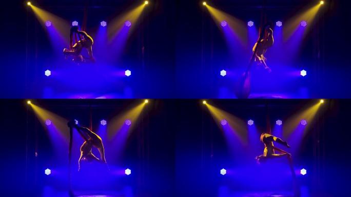女空中体操运动员在红色丝绸上旋转并缠绕。激动人心的杂技表演。深色烟熏背景和蓝色霓虹灯效果。慢动作