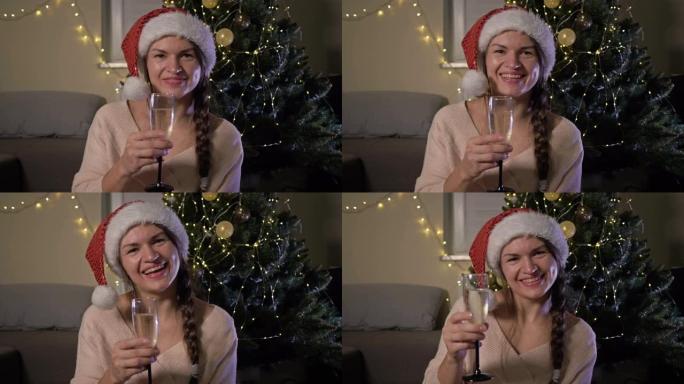 戴着圣诞老人帽子的美丽开朗的女人，拿着一杯香槟。在圣诞树的背景下。新年快乐，圣诞快乐
