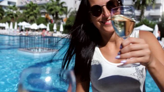 戴着太阳镜的微笑休闲女人在豪华棕榈度假村海滩POV拍摄时碰杯葡萄酒