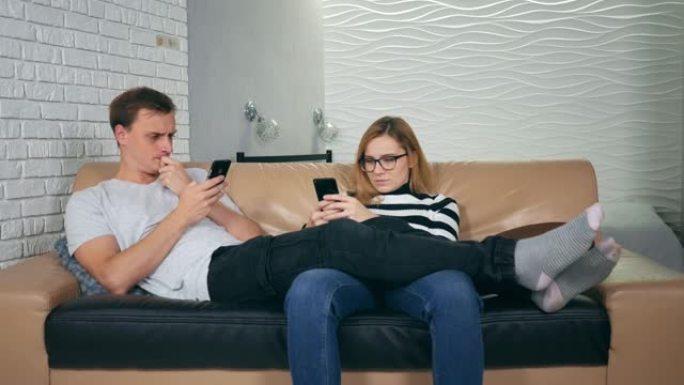 迷人的年轻夫妇坐在沙发上痴迷于智能手机。与手机相视对方为陌生人的夫妻，沟通问题和社交网络成瘾