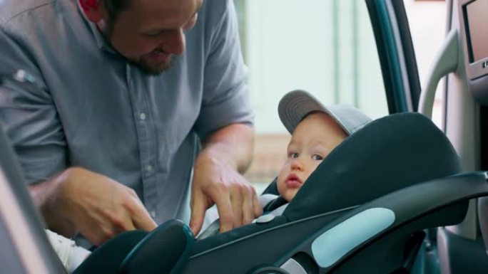 特写戴着帽子的男婴，坐在家用汽车内的婴儿汽车座椅上。门打开，他微笑着的大胡子父亲解开安全带，小心翼翼