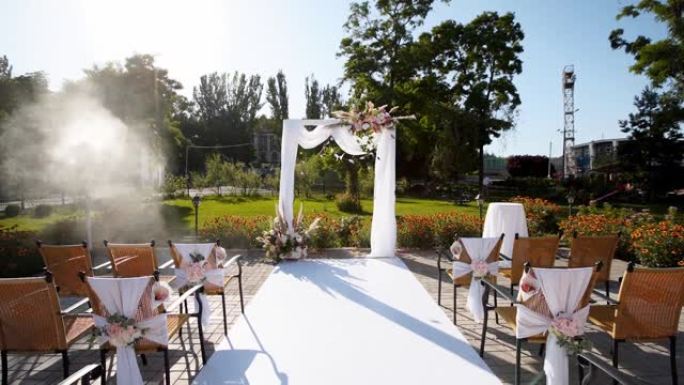 阳光明媚的夏日用鲜花装饰的婚礼拱门。椅子上的节日花卉装饰。玫瑰和羽毛草，白色纺织框架上有灯泡花环