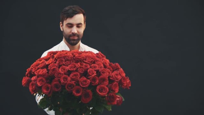 漂亮的男人站着，手里拿着一束大束红玫瑰，上面系着条纹，将其延伸到相机上，微笑着抬起眉毛。
