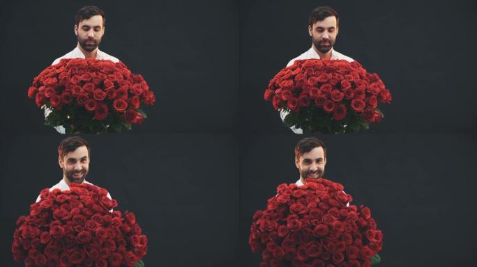漂亮的男人站着，手里拿着一束大束红玫瑰，上面系着条纹，将其延伸到相机上，微笑着抬起眉毛。