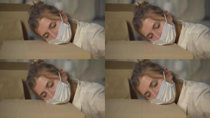 穿着Covid面罩睡在纸板箱上的疲惫的白人妇女的特写镜头。疲惫的年轻女商人的肖像，头躺在包裹上。冠状