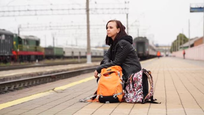 年轻女子蹲在站台上，等待火车。女乘客，背包坐在铁路平台上等待火车