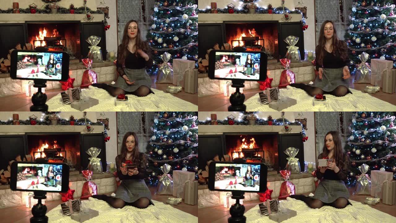 微笑的女人坐在燃烧的壁炉旁边令人惊叹的发光圣诞树背景上，用智能手机摄像头为在线观众录制视频。圣诞节假